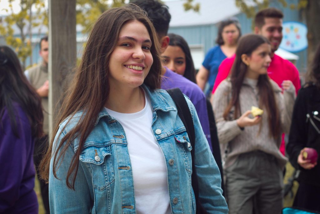 A young woman in a denim jacket smiles during an outdoor gathering with friends.